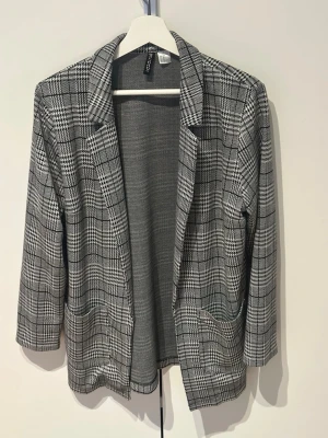 Grå rutig kavaj från H&M - Stilren grå kavaj med svartvitt rutmönster från H&M. Klassisk passform med två framfickor och smala slag. Materialet är mjukt och lätt, perfekt för en trendig look. 