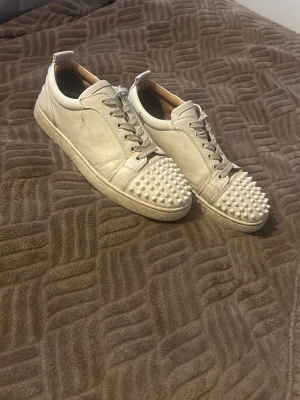 Vita sneakers med nitar från Christian Louboutin - Unika vita sneakers i läder från Christian Louboutin med coola nitar på tån och klassisk låg siluett. Skorna har vita snören, diskret logga och en chunky sula. Perfekta för dig som vill sticka ut med din stil. Dem är lite smutsiga nu men kan göra rent dem om man vill. Kan även sänka priset vid snabb affär 