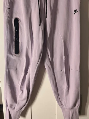 Ljuslila mjukisbyxor från Nike - Säljer ett par ljuslila mjukisbyxor från Nike med svarta detaljer och dragsko i midjan. Byxorna har en unik svart dragkedjeficka på ena benet och ribbade muddar vid bensluten. Perfekta för en avslappnad och sportig stil. De är knappast använda, och har legat orörda i garderoben väldigt länge.