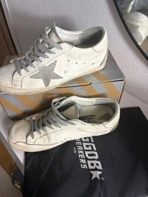 Vita Golden Goose sneakers med grå stjärna stl 45! - Säljer ett par vita Golden Goose sneakers med grå stjärna på sidan och grå skosnören. Skorna har en klassisk låg siluett och är tillverkade i läder med detaljer i mocka. Perfekta för dig som gillar en avslappnad och trendig stil. OBS! kolla sista bild för slitning i sulan