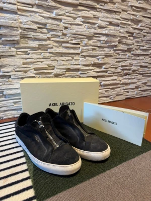 Svarta Axel Arigato sneakers med dragkedja - Svarta sneakers från Axel Arigato i mocka med cool dragkedja framtill istället för klassiska snören. Låg modell med vit sula och diskret logga på sidan. Perfekta för dig som gillar stilrena och unika detaljer i din outfit. Pris kan diskuteras!