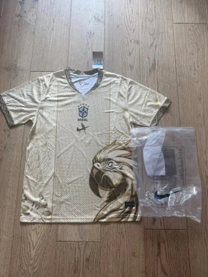 Guld Brasilien Nike fotbollströja - Snygg beige fotbollströja från Nike med Brasil-logga på bröstet och ett stort papegojmotiv framtill. Tröjan har korta ärmar, V-ringning och detaljerade mönster i guldtoner längs sidorna och ärmsluten. Tillverkad i lätt och ventilerande material, perfekt för träning eller match.