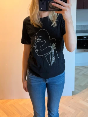 Svart t-shirt med ansiktsmotiv - Svart t-shirt med vitt ansiktsmotiv och dekorativ pärl-detalj som örhänge. Den vita färgen har flagnat lite.