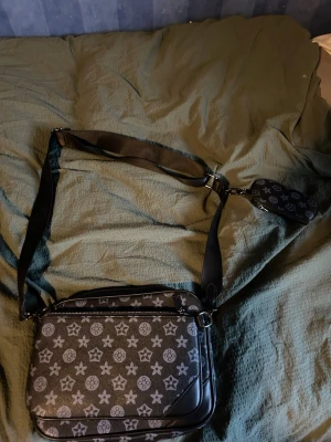 Axelväska svart - Snygg svart axelväska med klassiskt monogrammönster i grått. Väskan har justerbar axelrem, dragkedja och en mindre avtagbar ficka. Tillverkad i slitstarkt material med detaljer i läder. Perfekt för att bära dina viktigaste saker med stil.