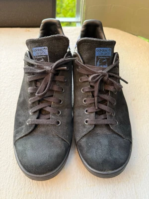Adidas Stan Smith svarta sneakers - Svarta Adidas Stan Smith sneakers i mocka med blå detalj och logga på hälen. Klassisk låg modell med snörning och diskret branding på plösen. Perfekta för en stilren och avslappnad look.