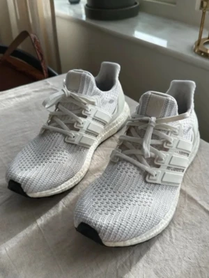 Vita Ultraboost  - Snygga vita Adidas Ultraboost sneakers med stickad ovandel i mesh och klassiska tre ränder på sidorna. Skorna har en bekväm och responsiv Boost-sula samt vita skosnören. Perfekta för en sportig och trendig look.