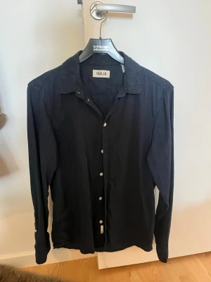 Marinblå skjorta från Solid - Klassisk marinblå skjorta från Solid med lång ärm och vita knappar framtill. Skjortan har en enkel, stilren design och är tillverkad i ett mjukt bomullsmaterial. Perfekt för dig som gillar en clean och tidlös look.