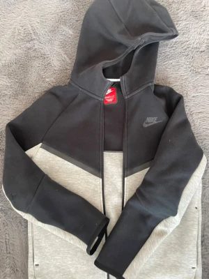 Nike tech fleece hoodie grå och svart - Snygg hoodie från Nike i svart och ljusgrått med dragkedja framtill och huva. Nike-logga på bröstet och liten logga bak. Tillverkad i mjukt sweatshirtmaterial, perfekt för en avslappnad streetwear-look.
