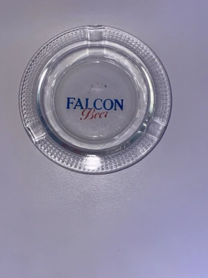 Falcon Beer askkopp i glas - Säljer en rund askkopp i genomskinligt glas med texten 'Falcon Beer' i blått och rött. Askkoppen har tre urtag för cigaretter och en prickig kant runt om. Perfekt för samlare eller som dekoration.