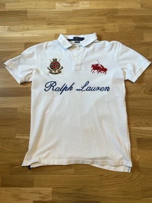 Vit pikétröja från Ralph Lauren - Vit pikétröja från Ralph Lauren med blå broderad logga och text på bröstet, röd broderad polospelare samt emblem. Korta ärmar med blått broderat nummer på ena ärmen. Klassisk krage och knappar framtill. Skick 9/10