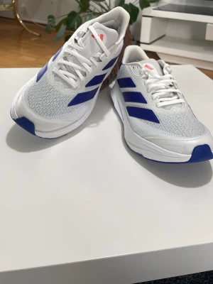 Vita och blå sneakers från Adidas  - Snygga sneakers från Adidas i vitt med blå detaljer och klassiska tre ränder på sidan. Ovandelen är i mesh för bra andningsförmåga och sulan är vit med blå inslag undertill. Perfekta för dig som gillar sportig och fräsch stil.