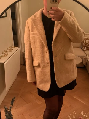 Beige ullkappa från Twist & Tango - Stilren beige kappa från Twist & Tango i ullmix med klassisk krage, två knappar framtill och tre fickor. Kappan har en mjuk, fluffig textur och är helfodrad med prickigt innerfoder. Perfekt för dig som vill ha en trendig och tidlös look. NYPRIS CA 3000