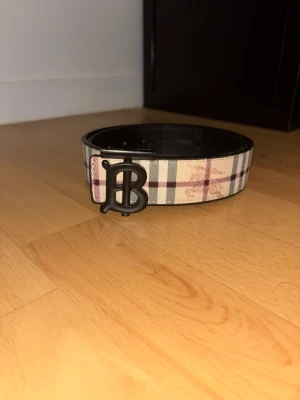 Rutmönstrat bälte från Burberry - Snyggt bälte från Burberry med klassiskt rutmönster i beige, svart och rött. Bältet har en stor metallspänne med Burberrys ikoniska B-logga och insida i svart skinn. Perfekt accessoar för att lyfta din outfit.