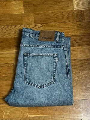 Blåa jeans från Woodbird - Säljer ett par klassiska blåa jeans från Woodbird i tvättad denim. Jeansen har raka ben, Snyggt läderpatch med logga bak i midjan. Perfekta för en avslappnad och trendig look. Stl W31 L34