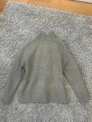 Grå stickad tröja från Zara - Mysig grå stickad tröja från Zara med hög ribbad krage och ribbade muddar vid ärmslut och nederkant. Tröjan har en enkel, klassisk design och är perfekt för kyliga dagar. Materialet känns mjukt och behagligt mot huden.