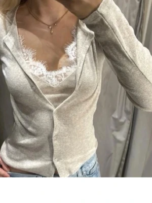 Beige kofta med spetsdetaljer - Snygg beige kofta med v-ringning och knäppning framtill. Under koftan syns ett linne med vit spetskant som ger en extra touch. Perfekt att matcha med jeans för en avslappnad men stilren look.