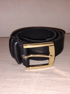 Svart bälte med guldfärgat spänne - Stilrent svart bälte med fyrkantigt guldfärgat spänne. Bältet är i klassisk design och passar perfekt till jeans eller byxor. Tillverkat i skinn för en tidlös look.