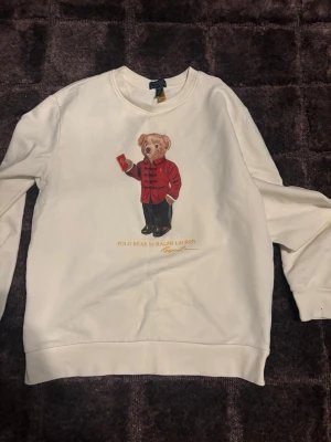 Vit sweatshirt Polo Bear Ralph Lauren - Vit sweatshirt från Ralph Lauren med det ikoniska Polo Bear-motivet på bröstet. Björnen har en röd jacka och håller i en kopp. Tröjan har rund halsringning, långa ärmar och ribbade muddar. Perfekt för dig som gillar klassisk streetwear med en twist.
