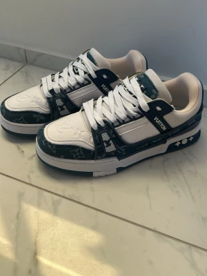 Louis Vuitton vita och blå sneakers - Snygga Louis Vuitton sneakers i vitt och blått med klassiskt LV-mönster. Skorna har vita snören, tjock sula och detaljer i svart och guld. Ikoniska LV-loggor syns både på sidan och bak. Tillverkade i läder och textil för en exklusiv känsla.