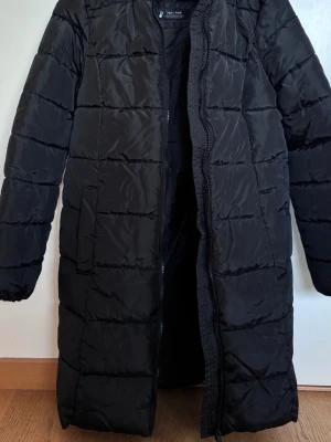 Svart lång dunjacka från DeFacto - Säljer en svart lång dunjacka från DeFacto med värmande HEAT PWR-teknologi. Jackan har quiltad design, dragkedja framtill och hög krage. Perfekt för kalla vinterdagar och riktigt skön att bära.