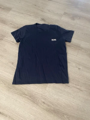 Mörkblå BOSS t-shirt med logga - Snygg mörkblå t-shirt från BOSS med liten vit logga på bröstet. Klassisk rund hals och korta ärmar. Tillverkad i mjuk bomull som känns skön mot huden. Perfekt till jeans eller shorts för en clean och enkel stil.