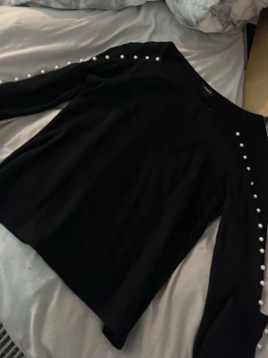 Svart långarmad tröja med pärldetaljer - Snygg svart tröja med rund hals och dekorativa pärlor längs ärmarna. Tröjan har en rak passform och långa ärmar. Perfekt för dig som gillar stilrena plagg med en twist.