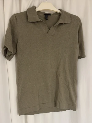 Oliv polotopp med krage - Snygg olivfärgad polotopp med klassisk krage och korta ärmar. Toppen har en avslappnad passform och är tillverkad i mjukt bomullsmaterial. Perfekt till jeans eller kjol för en enkel och stilren look.