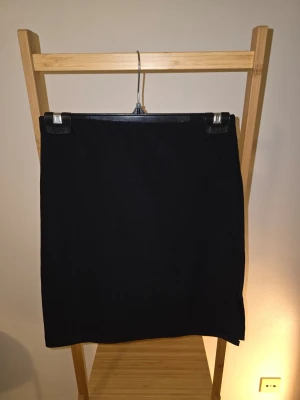 Svart kortkjol med slits - En enkel och stilren svart kortkjol med rak passform och liten slits på sidan. Kjolen är perfekt för att matcha med olika toppar och accessoarer. Materialet känns mjukt och följsamt. Oklart om storlek, den är lite stor på mig och jag är en S, skulle tippa på att den passar M.