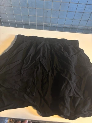 Svart kortkjol från Second Female - Säljer en svart kortkjol från Second Female med elastisk midja. Kjolen har en enkel och flowy design som gör den lätt att matcha med olika toppar. Perfekt för dig som gillar en stilren och bekväm look.