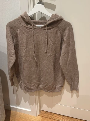 Beige stickad hoodie med huva - Mysig beige stickad hoodie med huva och dragsko. Tröjan har långa ärmar och ribbade muddar vid ärmslut och nederkant. Perfekt för dig som gillar en enkel och stilren look. Materialet känns mjukt och skönt mot huden.