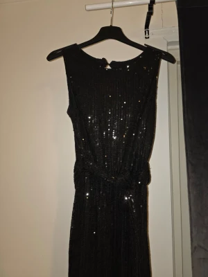 Svart glittrig jumpsuit med paljetter - Snygg svart jumpsuit med glittrande paljetter och vida ben. Ärmlös modell med rund halsringning och markerad midja. Perfekt för dig som vill sticka ut och glänsa lite extra. Materialet är stretchigt och följsamt för en bekväm passform.