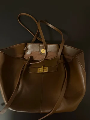 Brun bucket bag från Gina Tricot - Stilren brun bucket bag från Gina Tricot i slätt konstläder. Väskan har långa axelband, guldfärgat vridlås och magnetknapp. Invändigt finns en ficka med dragkedja och ett öppet fack. Perfekt för dig som gillar enkel men trendig design.