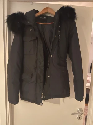 Svart pufferjacka med päls från Hollies - Säljer en svart pufferjacka från Hollies med stor fluffig päls på huvan. Jackan har dragkedja och knappar framtill, flera fickor och quiltad design. Perfekt för kalla dagar och har en snygg, modern look som funkar till många stilar.