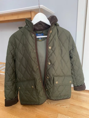 Olivgrön quiltad vindjacka Polo Ralph Lauren - Snygg olivgrön quiltad vindjacka från Polo Ralph Lauren med bruna detaljer, tryckknappar i guld, två stora fickor fram och avtagbar huva. Jackan har ribbade muddar och klassisk krage med brun manchester. Perfekt för dig som gillar stilren och tidlös design.