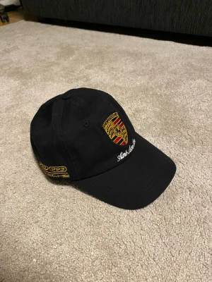 Aime Leon Dore Porsche Keps - Svart keps från Aime Leon Dore med broderad Porsche-logga framtill och gul text på sidan. Klassisk böjd skärm och justerbar passform bak. Materialet är bomull och kepsen har detaljer i vitt och gult. Perfekt för dig som gillar bilar och sportig stil.
