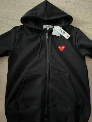 Svart hoodie Comme des Garçons Play - Svart hoodie från Comme des Garçons Play med dragkedja, huva och fickor framtill. Ikoniskt rött hjärta med ögon broderat på bröstet. Tillverkad i mjukt material som känns skönt mot huden. Perfekt för en avslappnad och trendig look.
