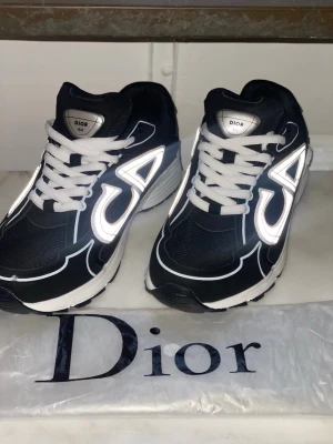 Dior b30 - Snygga svarta Dior sneakers med vita och reflekterande detaljer samt vita skosnören. Skorna har en chunky sula i vitt och svart och en sportig siluett. Materialet är en mix av mesh och syntet, vilket ger både komfort och stil. Perfekta för dig som vill sticka ut.