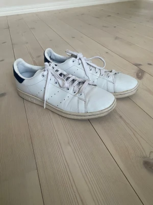 Adidas Stan Smith vita sneakers - Klassiska vita Adidas Stan Smith sneakers med mörkblå detalj på hälen och perforerade ränder på sidorna. Skorna har lågt skaft, vita snören och är tillverkade i läder. Perfekta för en clean och tidlös stil.