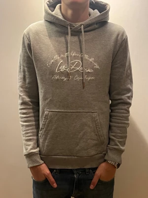 Grå hoodie Les Deux med tryck - Snygg grå hoodie från Les Deux med broderat vitt tryck på bröstet och texten 'Copenhagen'. Klassisk känguruficka framtill, ribbade muddar och justerbar huva med snören. Nypris 1300kr säljer pga att den är för liten. Storlek S, modellen är 181 cm lång 