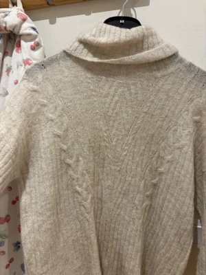 Beige polotröja med kabelstickning - Mysig beige stickad polotröja med kabelstickat mönster längs fram- och baksidan. Tröjan har ribbade detaljer och en hög, lös polokrage. Perfekt för kalla dagar och enkel att matcha med olika outfits.