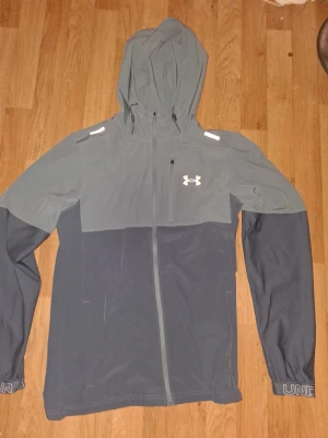 Grå vindjacka från Under Armour - Snygg tvåtonad grå vindjacka från Under Armour med huva och hel dragkedja. Jackan har logga på bröstet, diskreta reflexdetaljer på axlarna och textdetaljer på ärmsluten. Tillverkad i lätt och smidigt syntetmaterial, perfekt för blåsiga dagar.