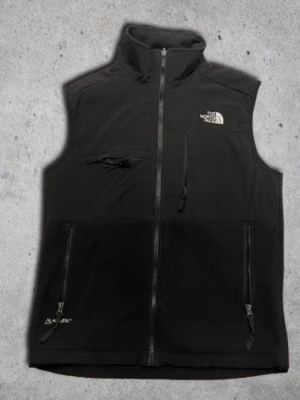 Svart fleeceväst från The North Face - Svart fleeceväst från The North Face med hög krage och dragkedja framtill. Västen har tre fickor med dragkedja och logga på bröstet. Perfekt för lager-på-lager och outdoor-vibe.