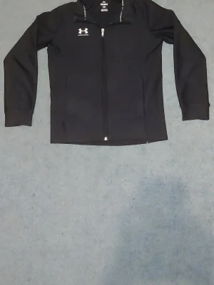 Svart ziptröja från Under Armour - Snygg svart ziptröja från Under Armour med hel dragkedja framtill och diskret logga på bröstet. Tröjan har lång ärm och hög krage, perfekt för lager på lager. Materialet är mjukt och stretchigt för extra komfort.