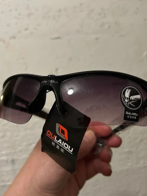 Svarta sportiga solglasögon Oulaiqu - Snygga svarta solglasögon från Oulaiqu med sportig design och halvbåge. Glasen är tonade i grått och bågarna är tillverkade i lätt syntetmaterial. Perfekta för dig som vill ha en cool och modern look i solen.