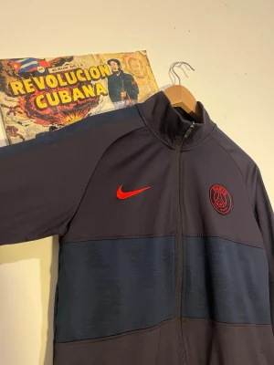 Mörkblå PSG träningsjacka Nike - Snygg mörkblå träningsjacka från Nike med PSG-logga och röd Nike swoosh på bröstet. Jackan har hel dragkedja, ståkrage och ribbade muddar. Perfekt för fotbollsträning eller chill. Materialet är troligtvis polyester för sportig känsla.