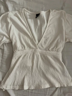 Vit omlottopp med v-ringning - Säljer en vit omlottopp från gina med djup v-ringning och lång ärm. Toppen har en lätt rynkad struktur och markerad midja som ger en snygg siluett. Perfekt att styla med jeans eller kjol för en trendig look.
