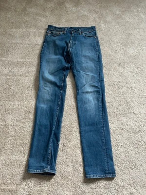 Blå raka jeans från Levi's med modellen 511 - Säljer ett par klassiska blå jeans från Levi's med raka ben och fem fickor. Jeansen har en snygg tvätt med slitningar och den ikoniska läderlappen bak i midjan. Perfekta för en avslappnad och tidlös stil.