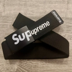 Svart Supreme bälte med logga - Supreme bälte i svart med tydlig vit logga på metallspännet. Bältet är gjort i slitstarkt tygmaterial och har en clean, streetwear-inspirerad look. Perfekt accessoar för att lyfta din outfit med en ikonisk detalj.