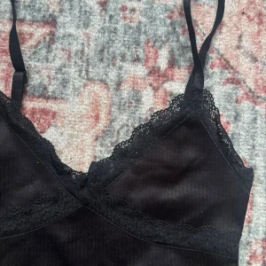 Svart spetslinnetopp med smala band - Snygg svart linnetopp med tunna axelband och spetsdetaljer längs urringningen och under bysten. Toppen har en croppad passform och är tillverkad i ett mjukt, stretchigt material som känns bekvämt mot huden. 