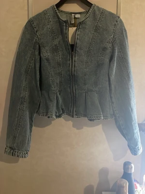 Blå jeansjacka med peplum - Trendig blå jeansjacka från H&M med peplumdetalj nedtill och dragkedja framtill. Jackan har långa ärmar och markerad midja som ger en snygg siluett. Perfekt för dig som vill ha en unik twist på den klassiska jeansjackan.